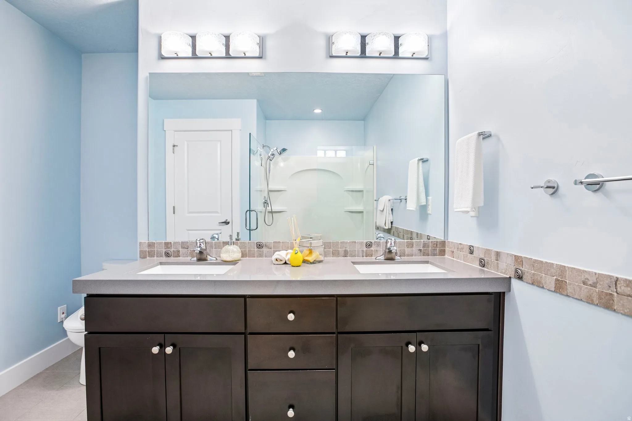 Dual integrated sinks set in a gray countertop, dark wood-finish vanity with drawers and cabinet storage, large vanity mirror, and recessed tile backsplash