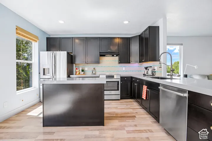 Modern kitchen featuring dark cabinetry with stainless steel appliances, a large waterfall-edge island with seating, white subway tile backsplash, quartz countertops, and wood-finish flooring throughout the open layout