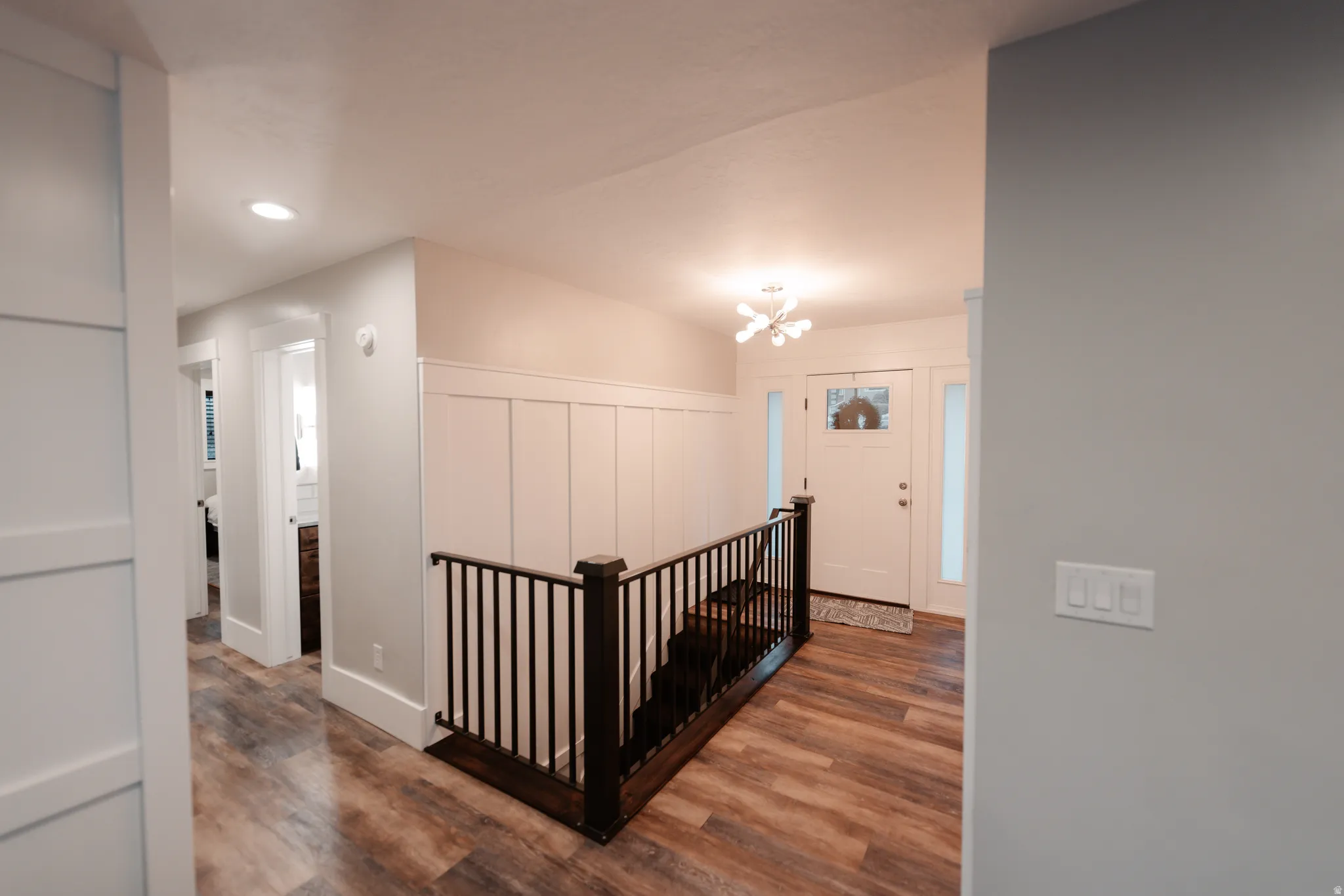 Entryway featuring wood-finish flooring, white paneled wainscoting, a contemporary chandelier, a white entry door with sidelights, and dark wood newel post and balusters