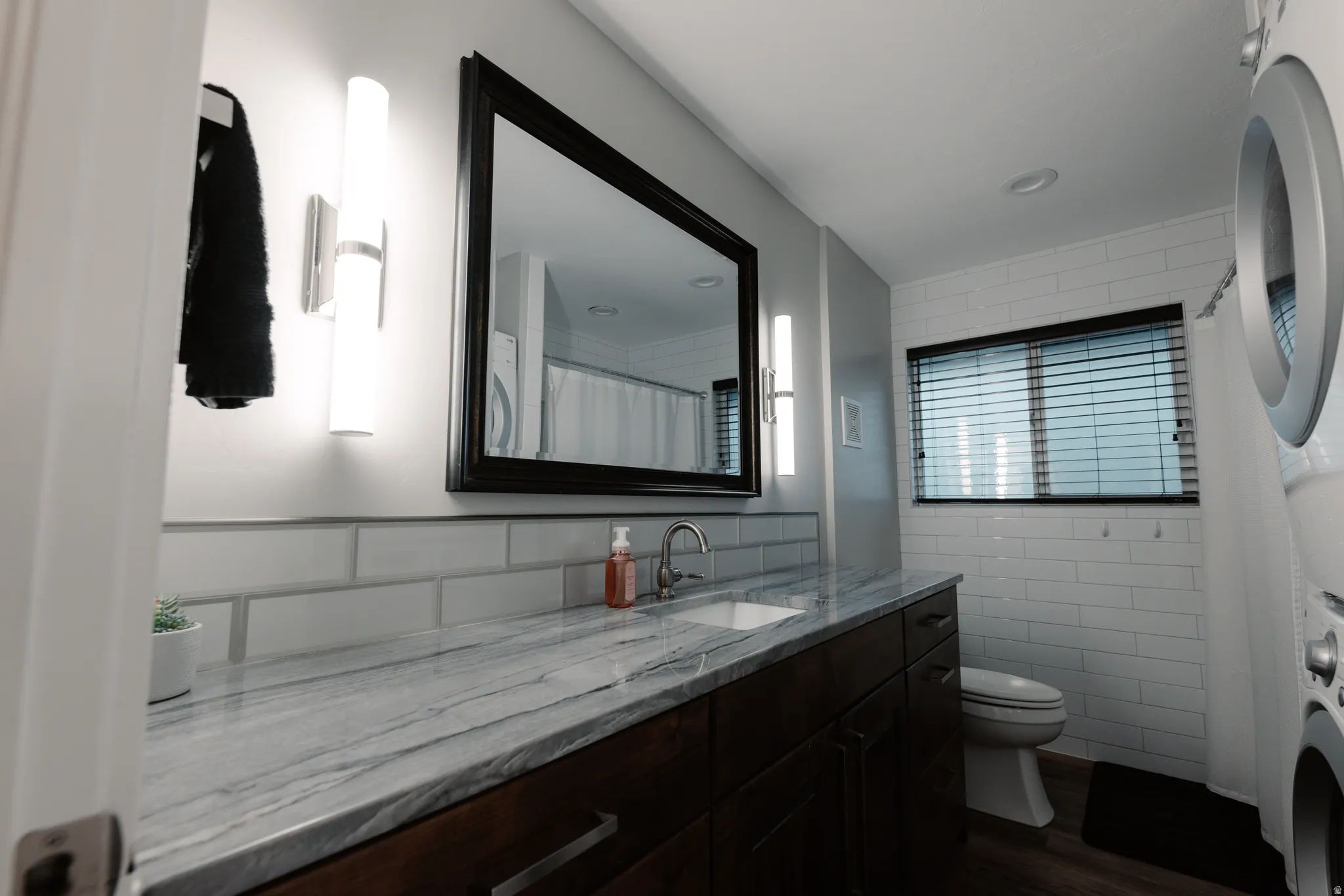 Modern bathroom featuring a double vanity with marble-look countertop, dual integrated lighting, black-framed mirror, subway tile backsplash, and in-unit laundry appliances