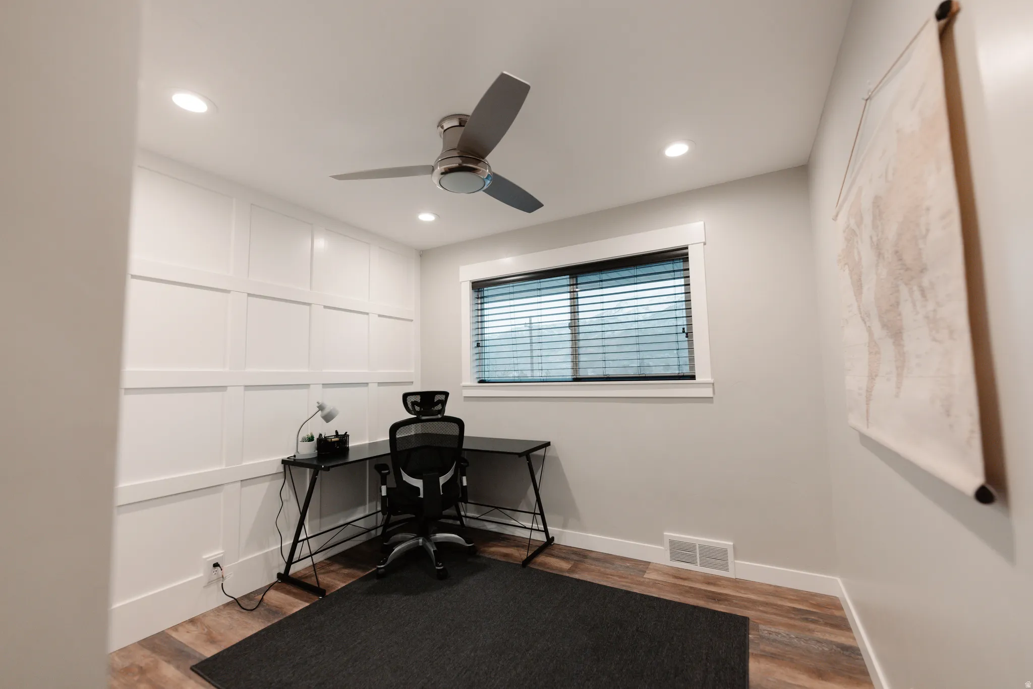 Dedicated home office featuring white shaker-style wall paneling, engineered wood-finish flooring with contrasting border detail, ceiling-mounted fan with integrated lighting, horizontal blinds, and neutral wall-mounted shelving