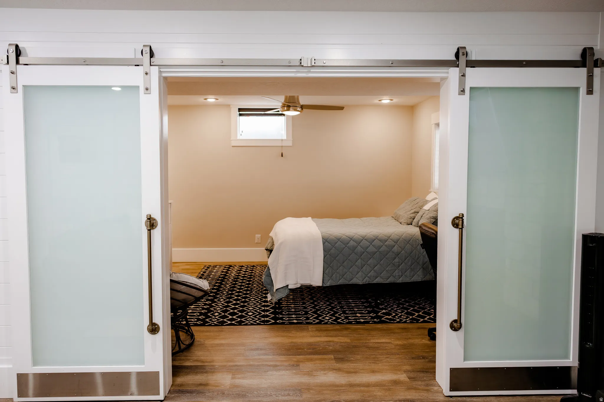 Spacious basement bedroom suite with barn-style sliding doors, wood-finish flooring, warm neutral wall finishes, and recessed ceiling lighting