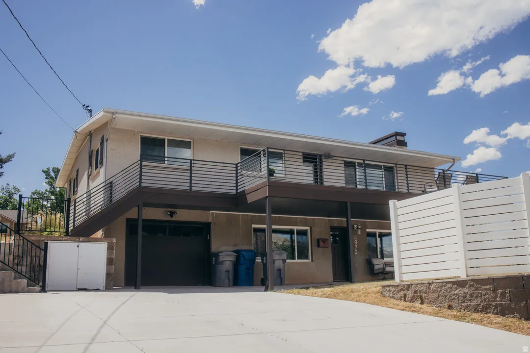 Modern two-story residence featuring a cantilevered upper level with horizontal metal railings, expansive concrete driveway, and ground-level garage