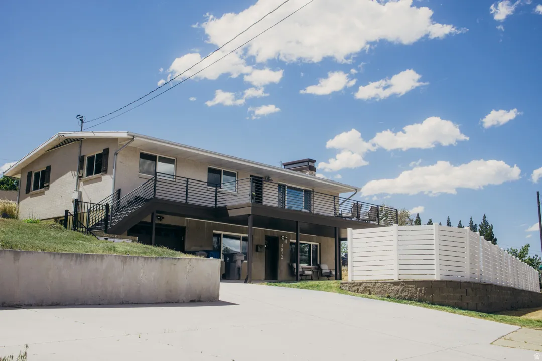 Modern split-level home featuring a contemporary black and wood exterior, expansive concrete driveway, horizontal slatted railings, large multi-pane windows, and mature evergreen landscaping along the perimeter