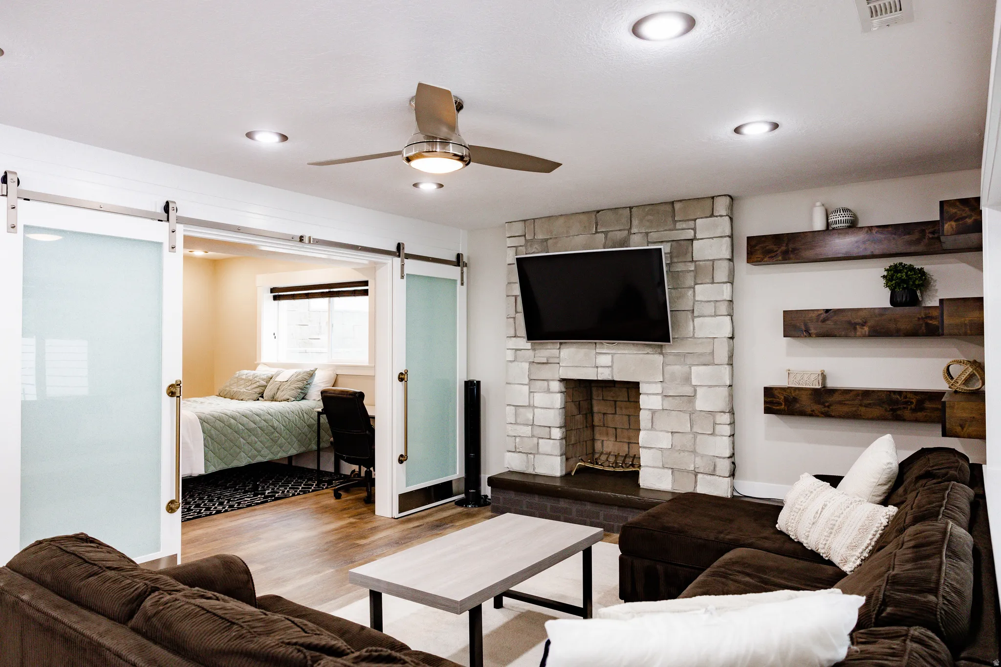 Basement Stone-clad fireplace with hearth, recessed lighting, wood-finish flooring, ceiling fan, and sliding frosted-glass barn doors