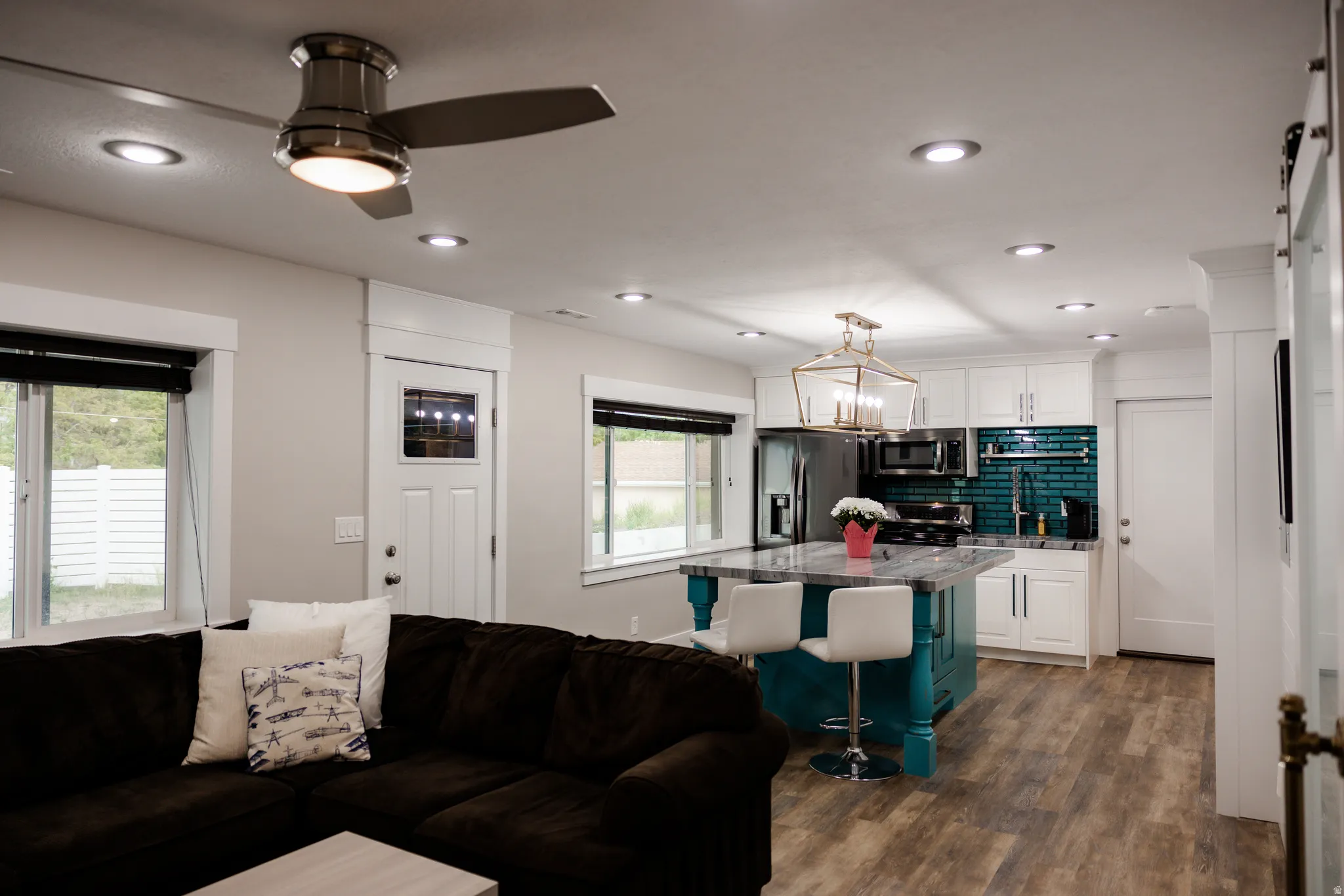 Open-concept living and kitchen area featuring white cabinetry with black hardware, a teal-finished kitchen island with seating, stainless steel appliances, black subway tile backsplash, wood-finish flooring throughout, recessed lighting, and a ceiling fa