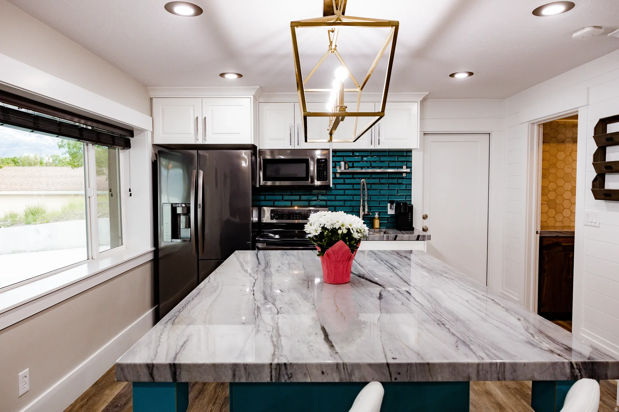Basement Modern kitchen featuring a waterfall-edge island with veined stone countertops, black stainless steel appliances, white cabinetry with brushed metal hardware, teal subway tile backsplash, recessed and pendant lighting, and large windows with natu