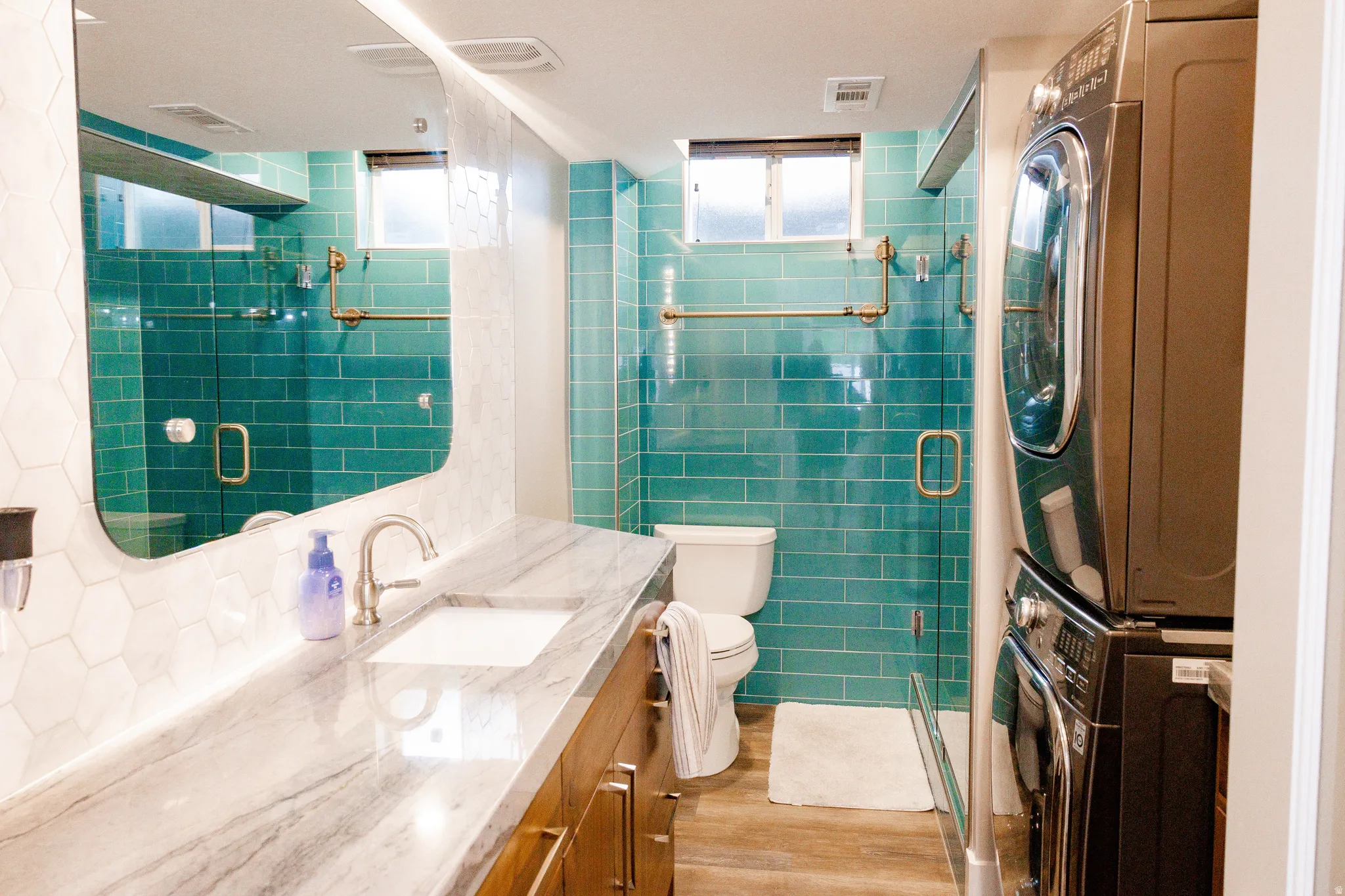 Spa-inspired primary bathroom featuring turquoise subway tile walls with brass hardware accents, a double vanity with white marble countertops, and a frameless glass-enclosed shower in basement