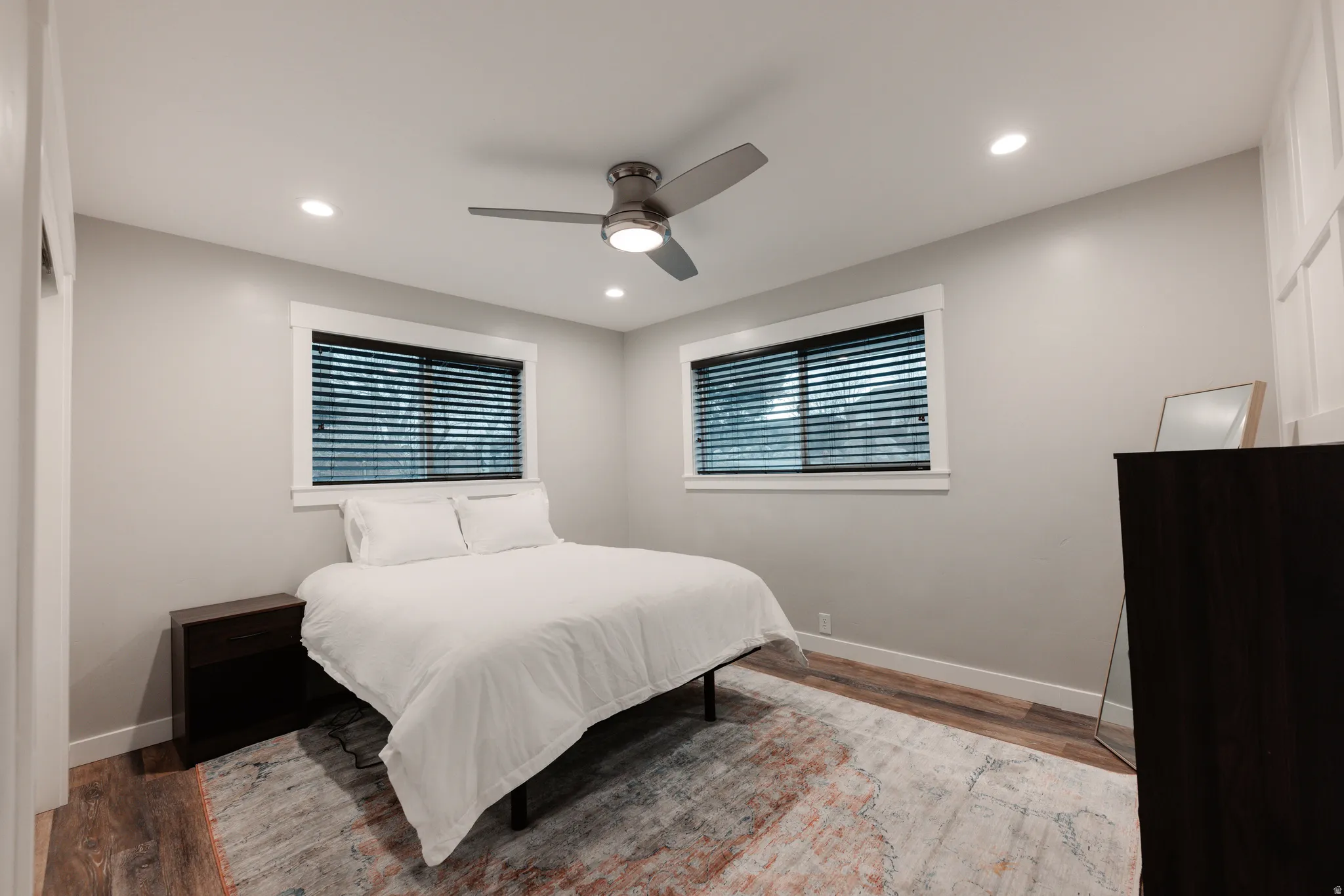 Bright secondary bedroom showcasing recessed ceiling lighting, ceiling fan with integrated light fixture, dual windows with horizontal blinds providing natural light, wood-finish flooring, and geometric area rug