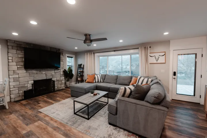 Spacious living room featuring a sectional layout anchored by a fireplace with brick surround, wall-mounted flat-screen television, recessed ceiling lighting, ceiling fan, and expansive windows with glass door access to outdoor space