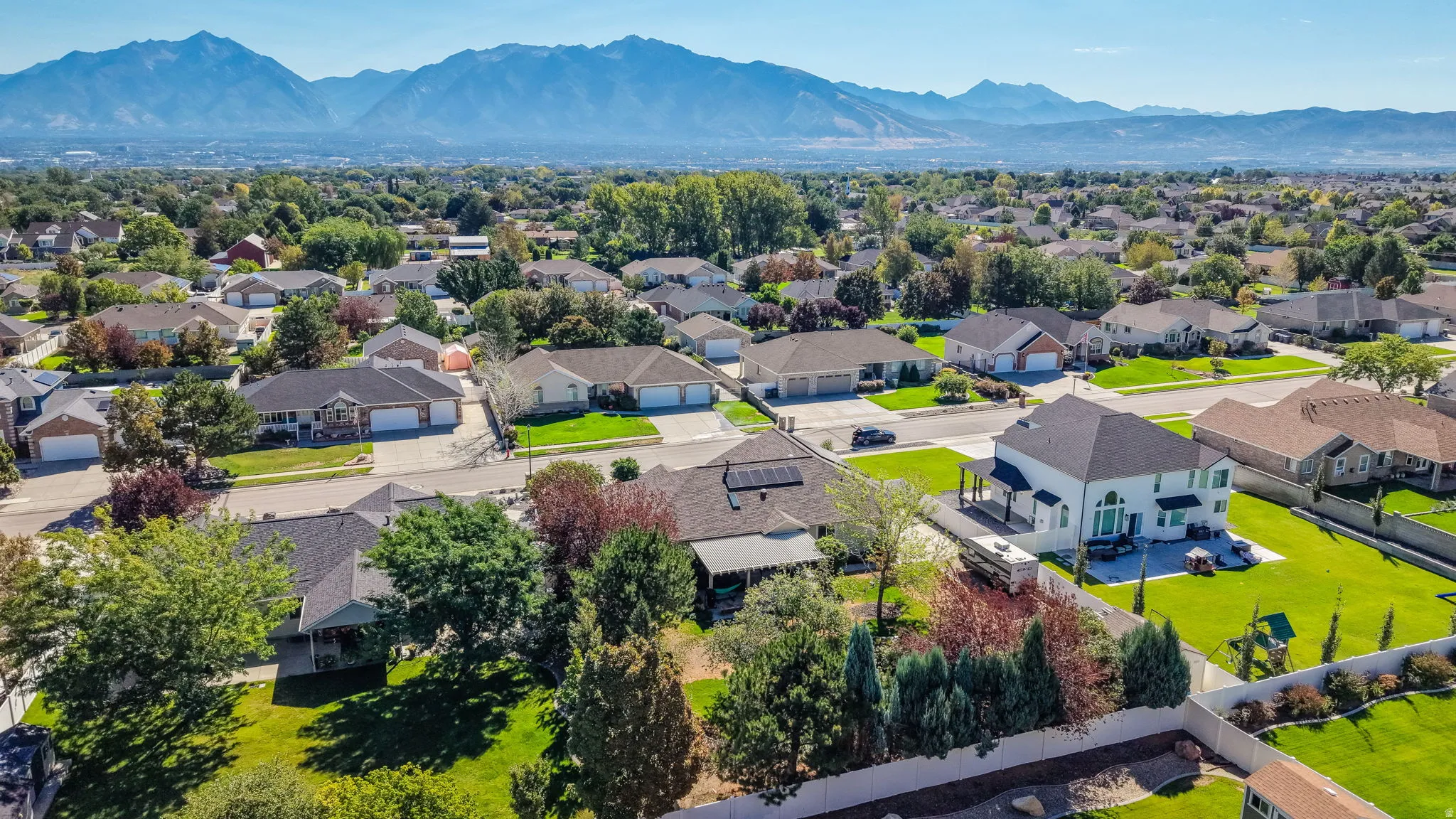 Expansive residential landscape with distant mountain ranges, featuring a two-story residence with a multi-car driveway, a large manicured lawn, and mature trees