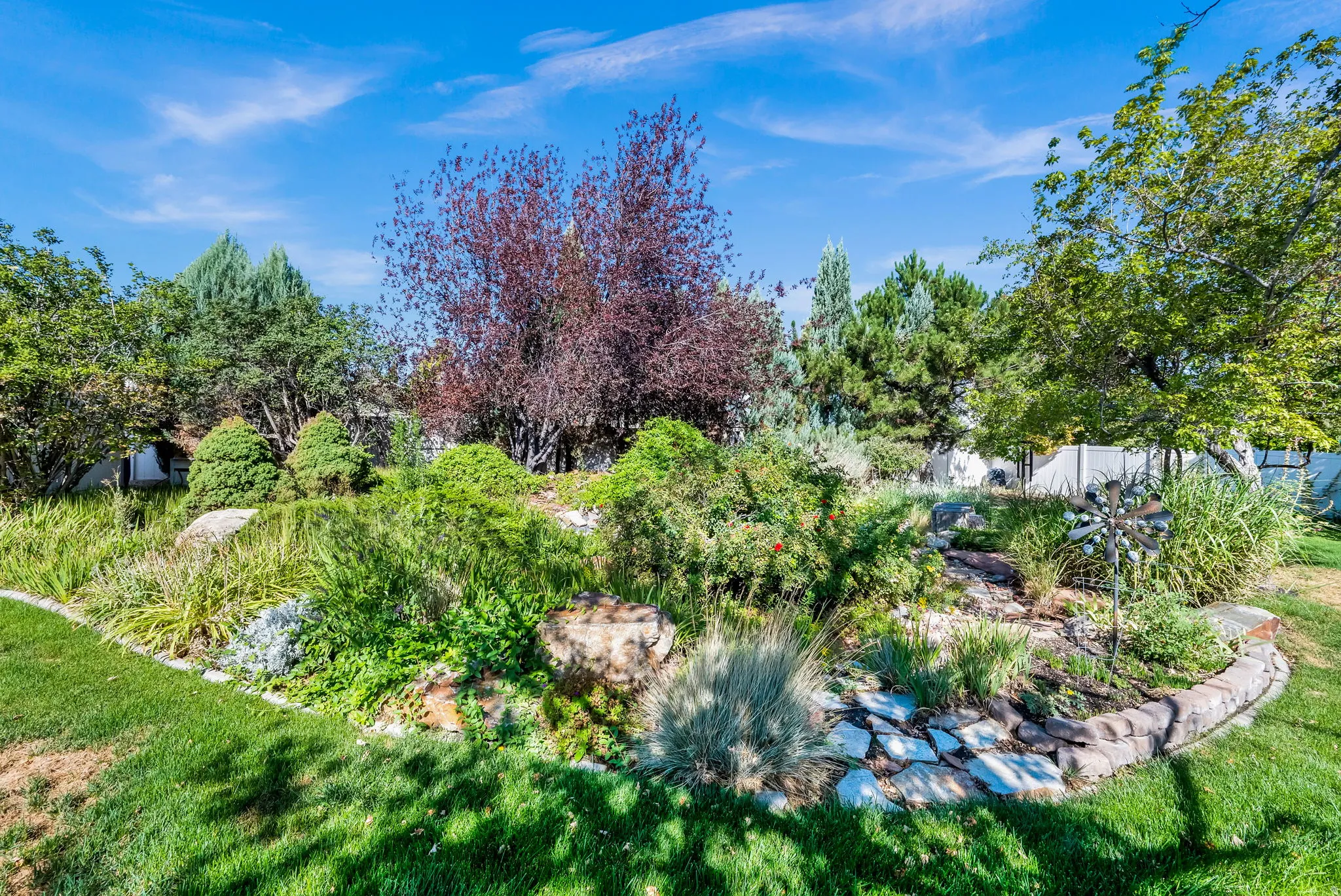 Mature landscaped grounds featuring lush green lawn, curved stone hardscaping, mixed plantings including ornamental trees with seasonal foliage, and established shade trees throughout the property