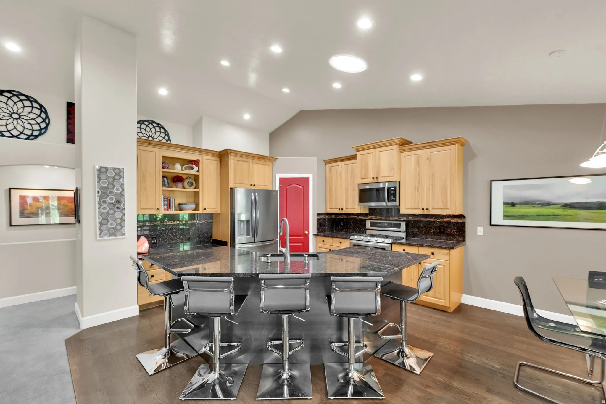 Open-concept kitchen featuring an angular island with seating, stainless steel appliances, wood cabinetry, and dark countertops