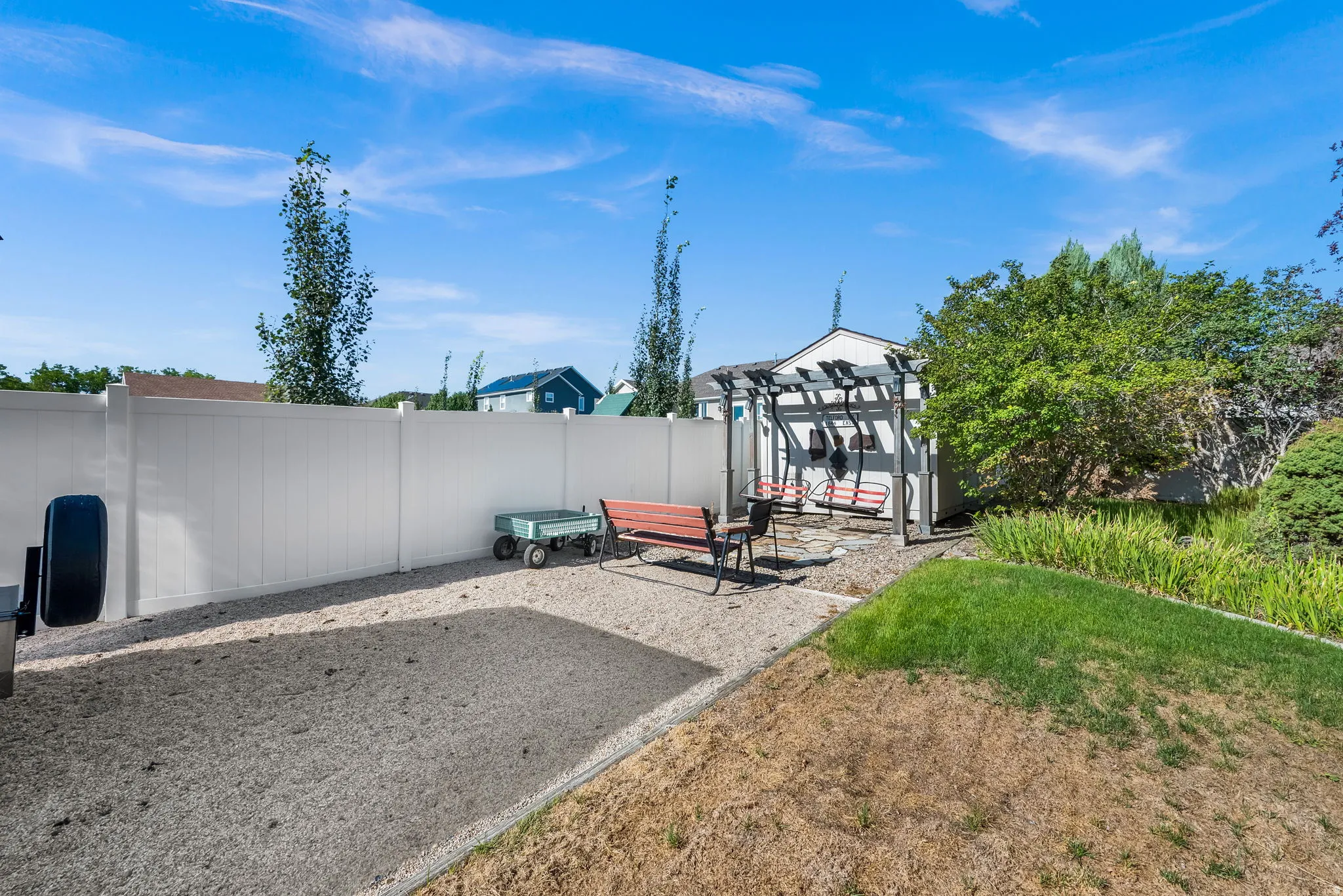 Backyard retreat featuring a gravel patio with modern pergola structure, mature landscaping including ornamental trees and shrubs, and a detached storage building with metal siding