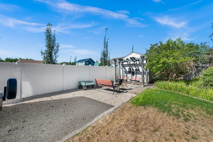 Backyard retreat featuring a gravel patio with modern pergola structure, mature landscaping including ornamental trees and shrubs, and a detached storage building with metal siding