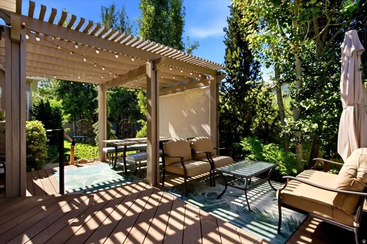 The custom backyard retreat features a large deck with a pergola cover, patio lighting, and mature trees.