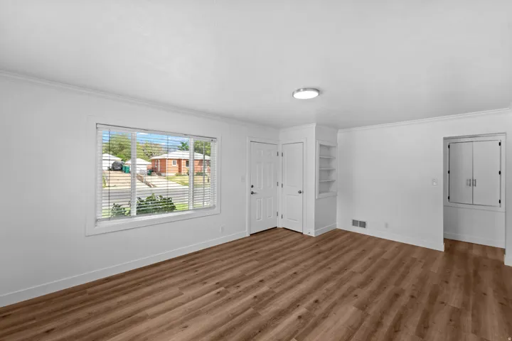 Spacious room featuring wood-finish flooring, a large window with blinds, a built-in shelving unit, and a flush-mount ceiling light