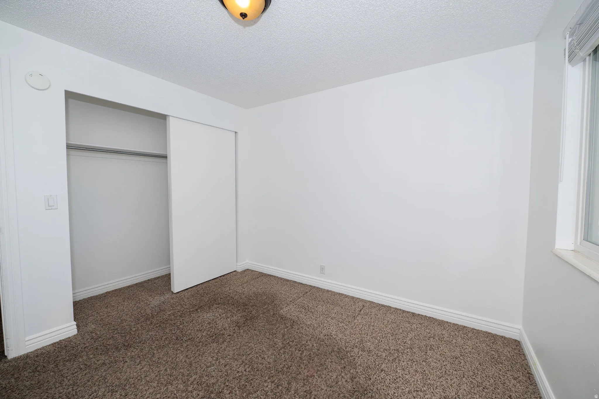 Light brown carpet flooring, white walls, built-in closet with sliding doors, single window, and a ceiling-mounted light fixture