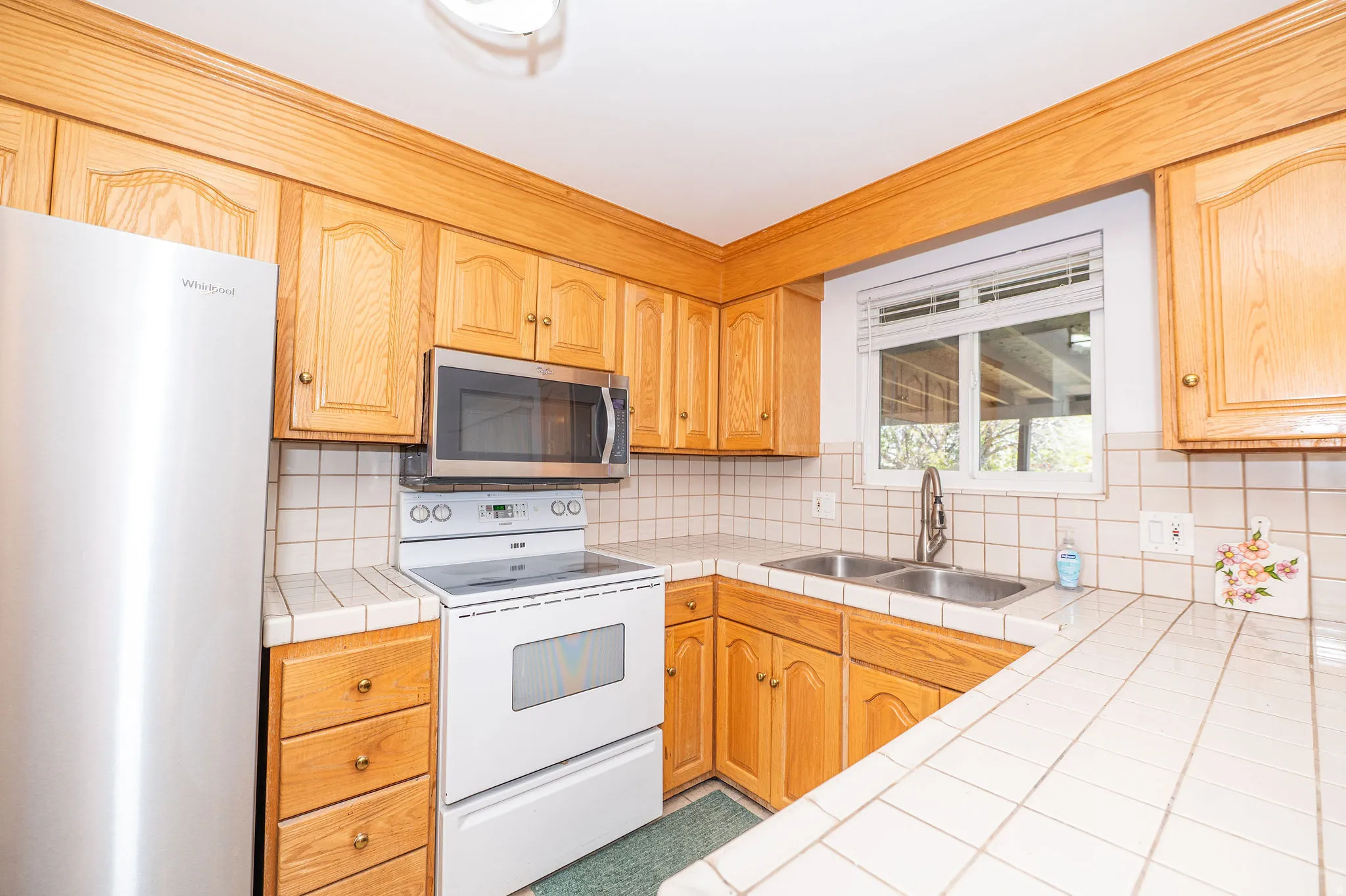 Kitchen featuring wood cabinetry, stainless steel refrigerator, white electric range with microwave, tile countertops, and a double basin stainless steel sink