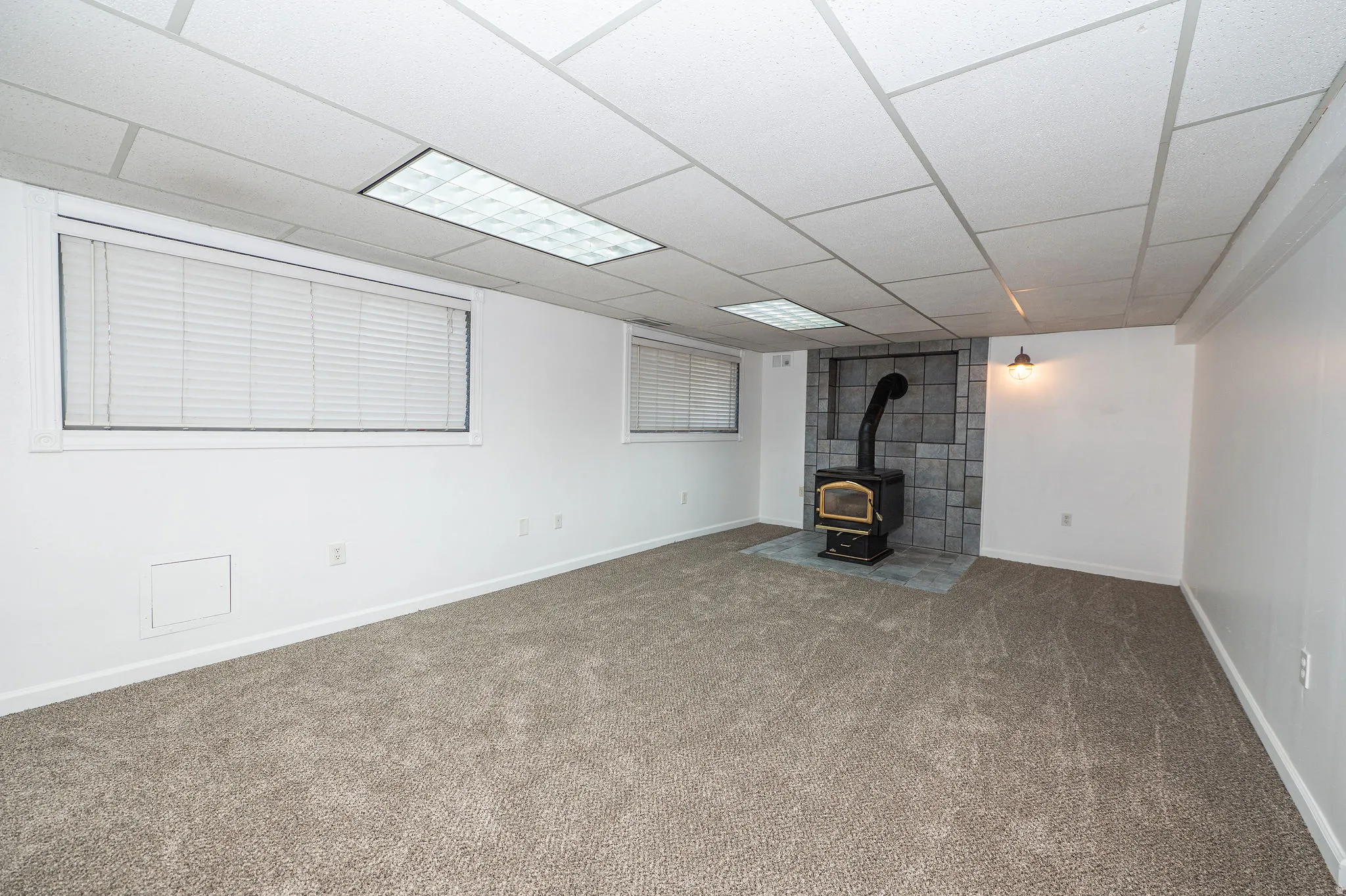 Carpeted room featuring a wood-burning stove with a tile surround and hearth, multiple windows, and a drop ceiling with integrated lighting