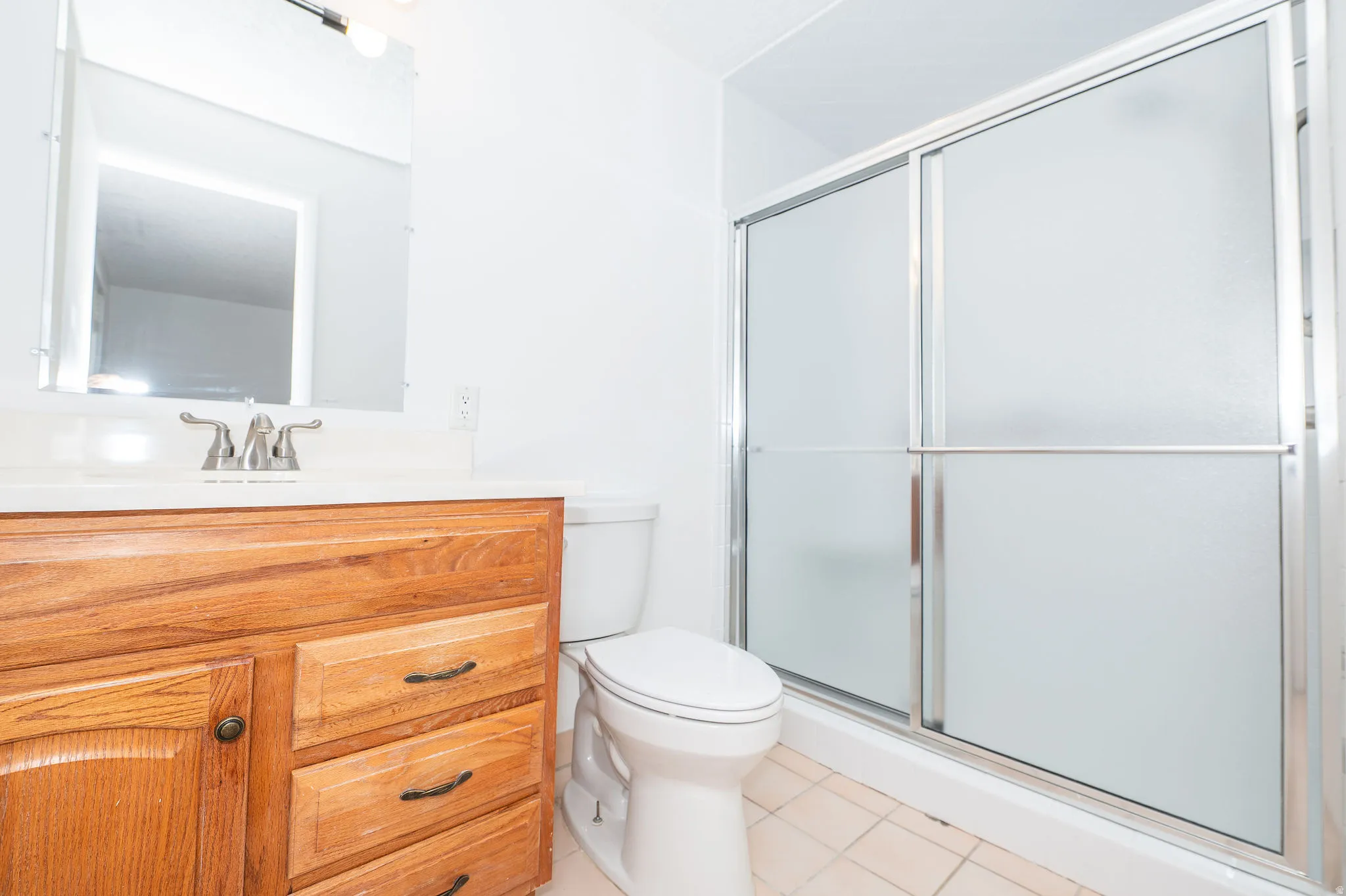 White vanity countertop, wood-finish cabinetry, chrome faucet, and frosted glass shower enclosure