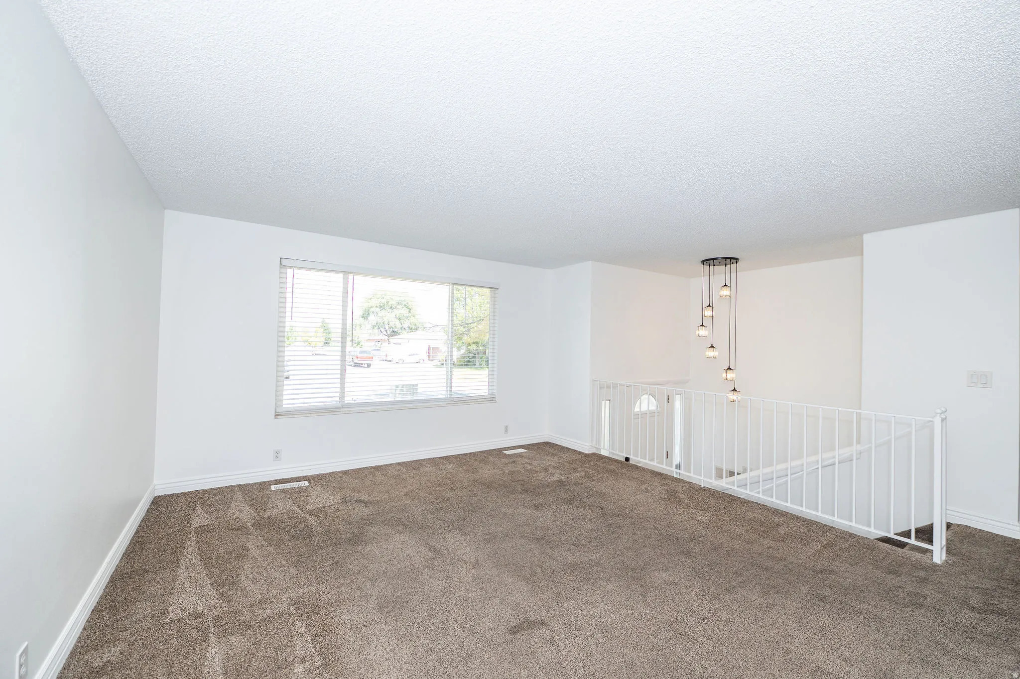 White walls, brown carpet, window with horizontal blinds, white railing, and a multi-pendant light fixture