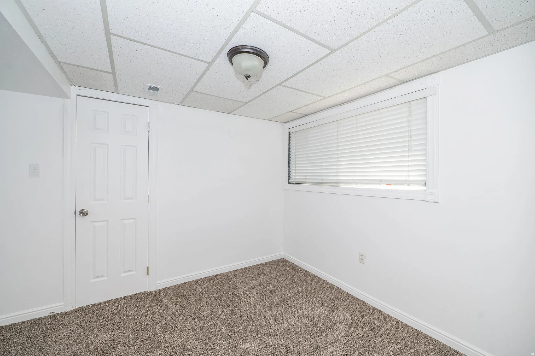 White painted walls, drop ceiling, flush mount light fixture, six-panel interior door, and window with white blinds