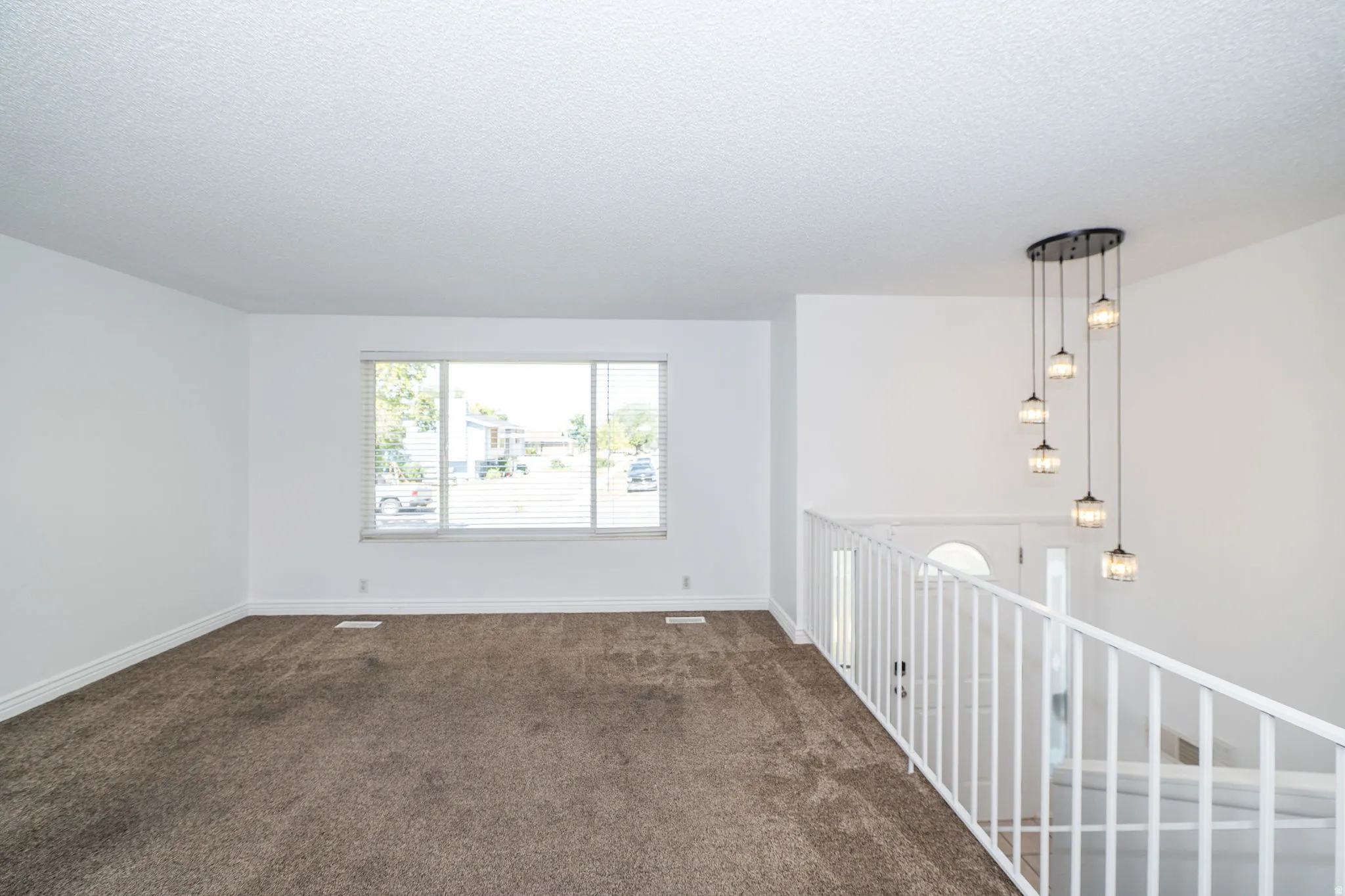 Plush carpet flooring, large window with blinds, white railing, and a multi-pendant light fixture