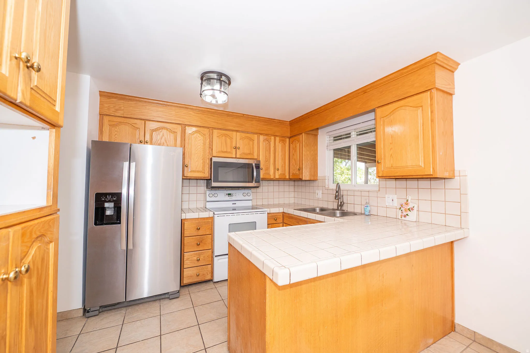 Kitchen featuring wood cabinetry, stainless steel refrigerator, tile countertops, tile backsplash, and a window above the sink