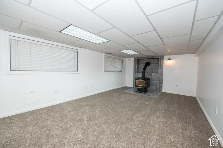Carpeted room featuring a wood-burning stove with a tile surround and hearth, multiple windows, and a drop ceiling with integrated lighting