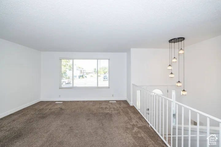 Plush carpet flooring, large window with blinds, white railing, and a multi-pendant light fixture