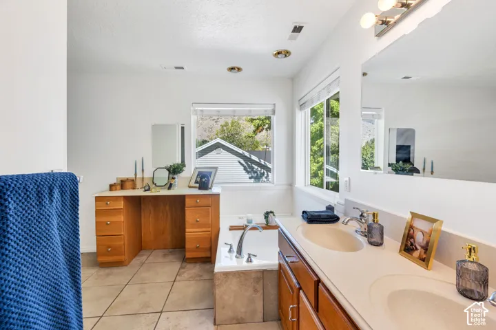 Dual vanity with integrated sinks, wood-finish cabinetry, built-in soaking tub, tile flooring, and integrated windows