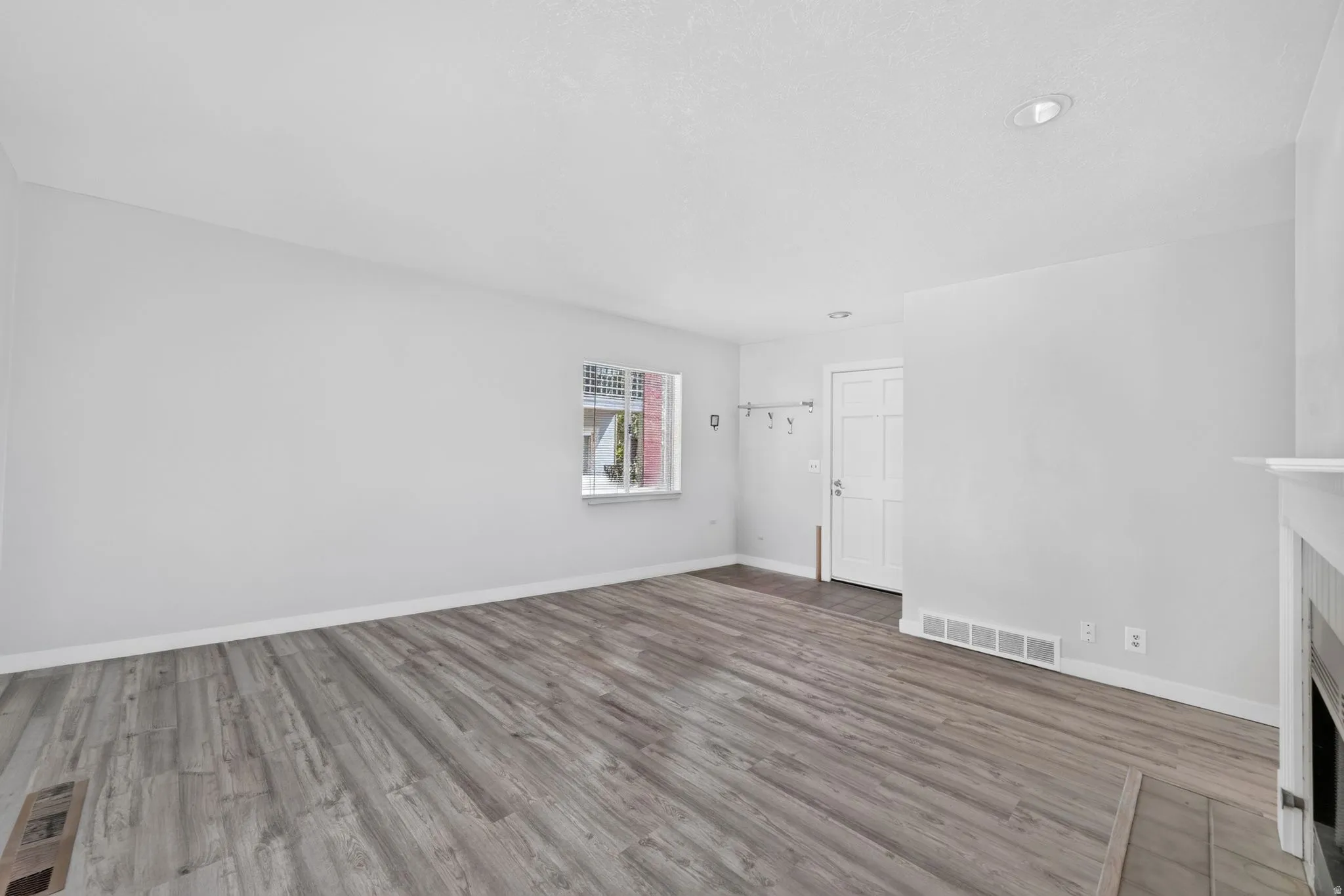 Spacious room featuring wood-finish flooring, a white paneled entry door, a window with grid detailing, and a built-in fireplace with a white mantel