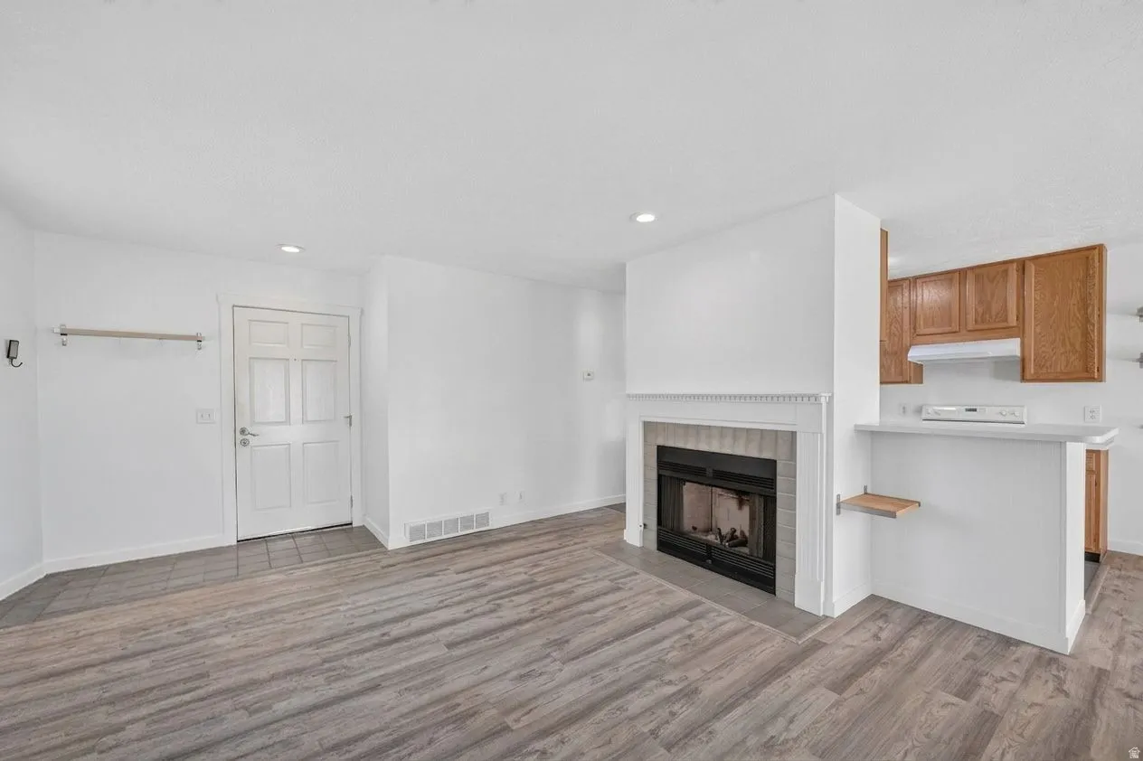 Wood-finish flooring, white walls, built-in fireplace with a mantel, white kitchen cabinetry with a built-in oven range, and an attached breakfast bar