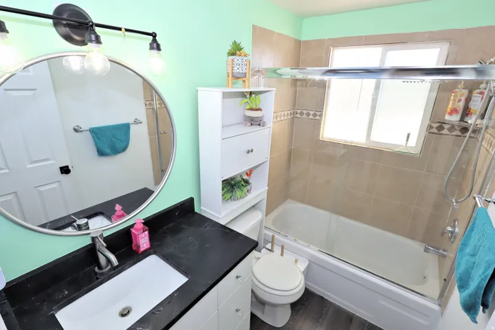 Bathroom featuring a vanity with a dark countertop and undermount sink, round mirror, built-in tub with tile surround, and glass shower enclosure