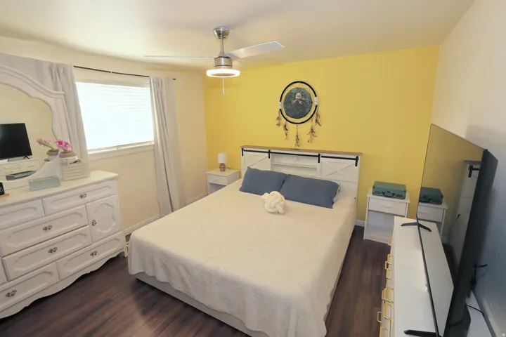 Wood-finish flooring, ceiling fan with integrated lighting, window with blinds and drapes, white dresser with mirror