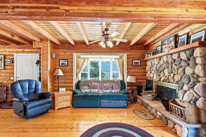 Rustic log cabin interior showcasing exposed beam ceiling with ceiling fan, hardwood flooring throughout, and a striking stone fireplace with substantial stacked-stone surround