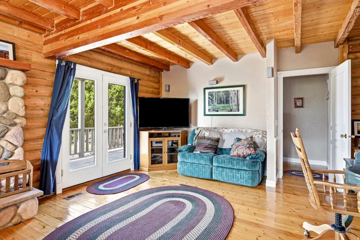 Exposed wood beam ceiling, wood-finish flooring, stone fireplace with hearth, and double French doors opening to a balcony