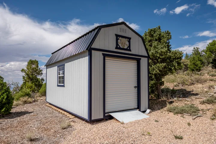 Detached shed featuring a gambrel roof, corrugated metal roofing, an overhead garage door with a ramp, and a single window