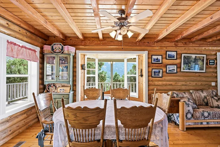 Log construction walls, exposed wood beam ceiling, wood-finish flooring, ceiling fan with integrated lighting, and multiple windows