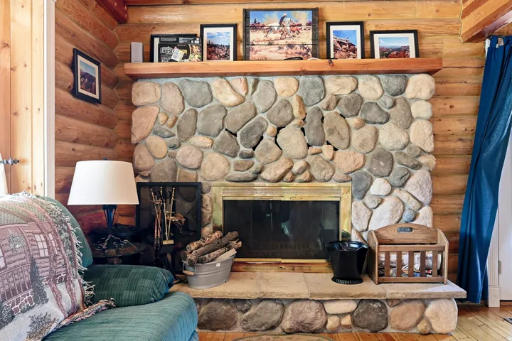 Rustic log cabin interior featuring a striking stone fireplace with substantial stacked-stone surround, wood-beam ceiling construction, and wood-finish flooring throughout