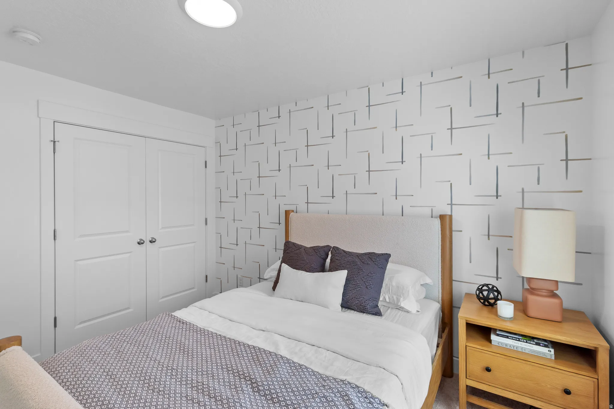 Recessed ceiling lighting, patterned accent wall, light wood-finish nightstand, light wood-finish bed frame, and crisp white bi-fold closet doors