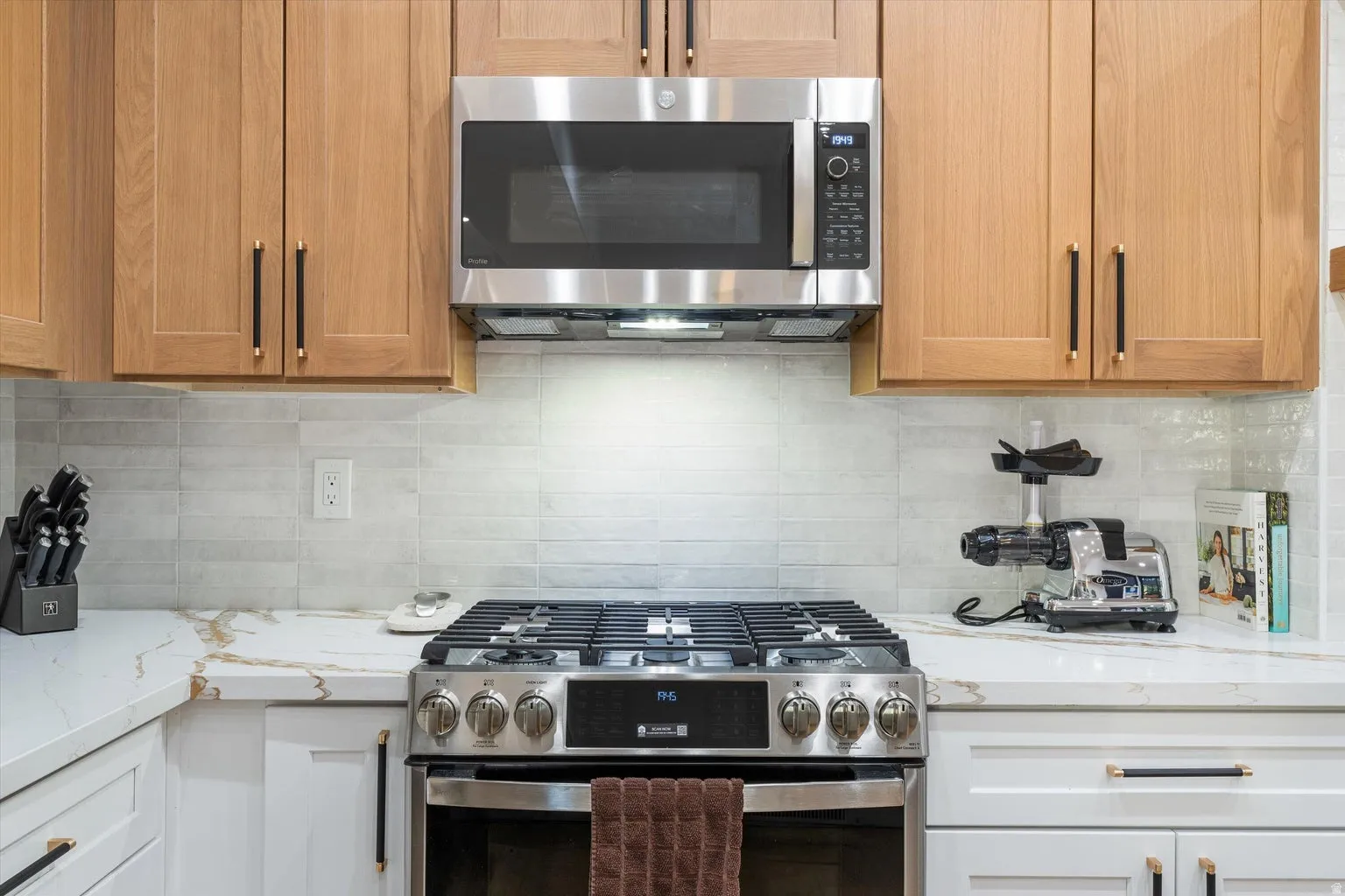Stainless steel gas range, integrated microwave, wood-finish cabinetry, white countertops with veining, and a light-toned tile backsplash