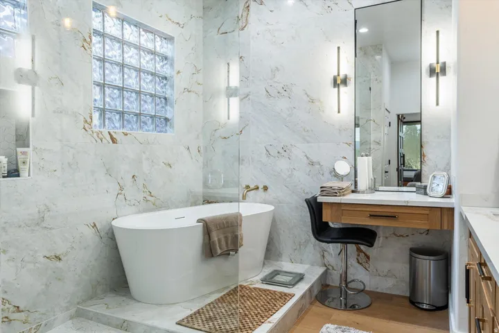 Freestanding soaking tub with gold-finish fixtures, marble-look wall tiling, glass block window, built-in vanity with wood-finish cabinetry, and full-height mirror