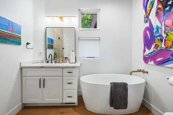 Freestanding oval soaking tub with wall-mounted brass-finish faucet, white vanity with stone countertop, rectangular vanity mirror, and wood-finish flooring