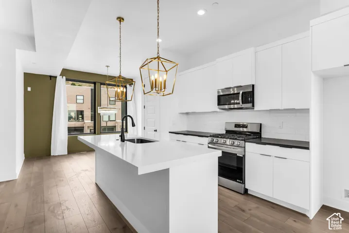 Modern kitchen featuring white cabinetry, a large waterfall-edge island with white quartz countertops, stainless steel gas range and built-in appliances, pendant lighting with brass fixtures, and wide-plank wood-finish flooring throughout the open-concept