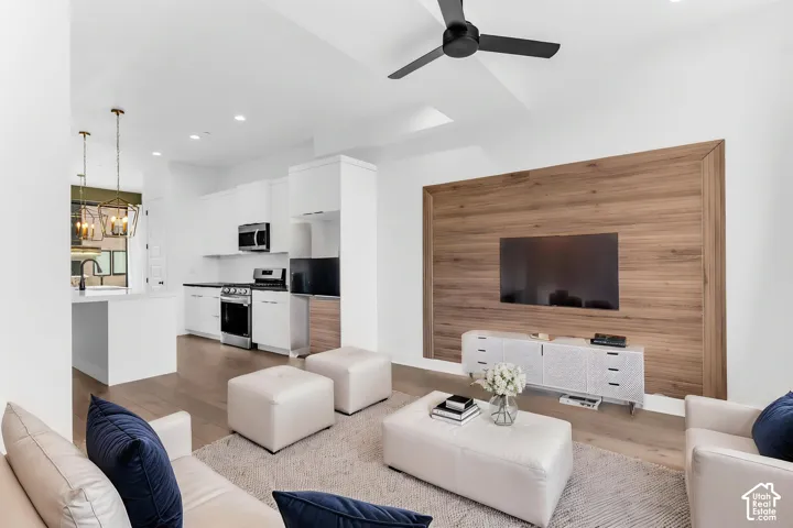 AI Staged - Open-concept living area featuring a wall-mounted entertainment display, white cabinetry with integrated storage, wood-finish accent wall, recessed and pendant lighting, ceiling fan, and a modern kitchen with stainless steel appliances and whi