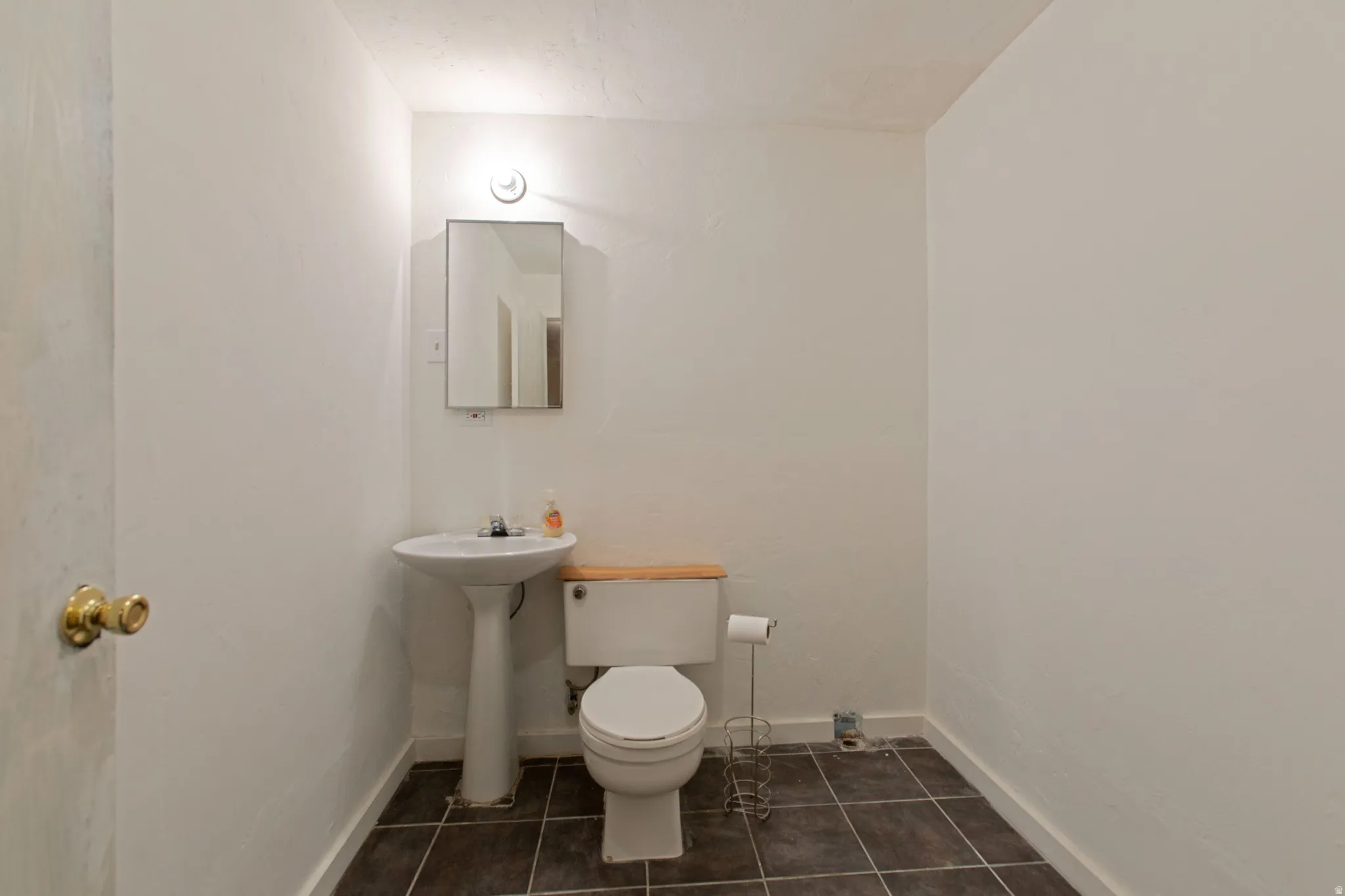 Pedestal sink, wall-mounted medicine cabinet, wall-mounted light fixture, white toilet with wooden lid, and dark tile flooring