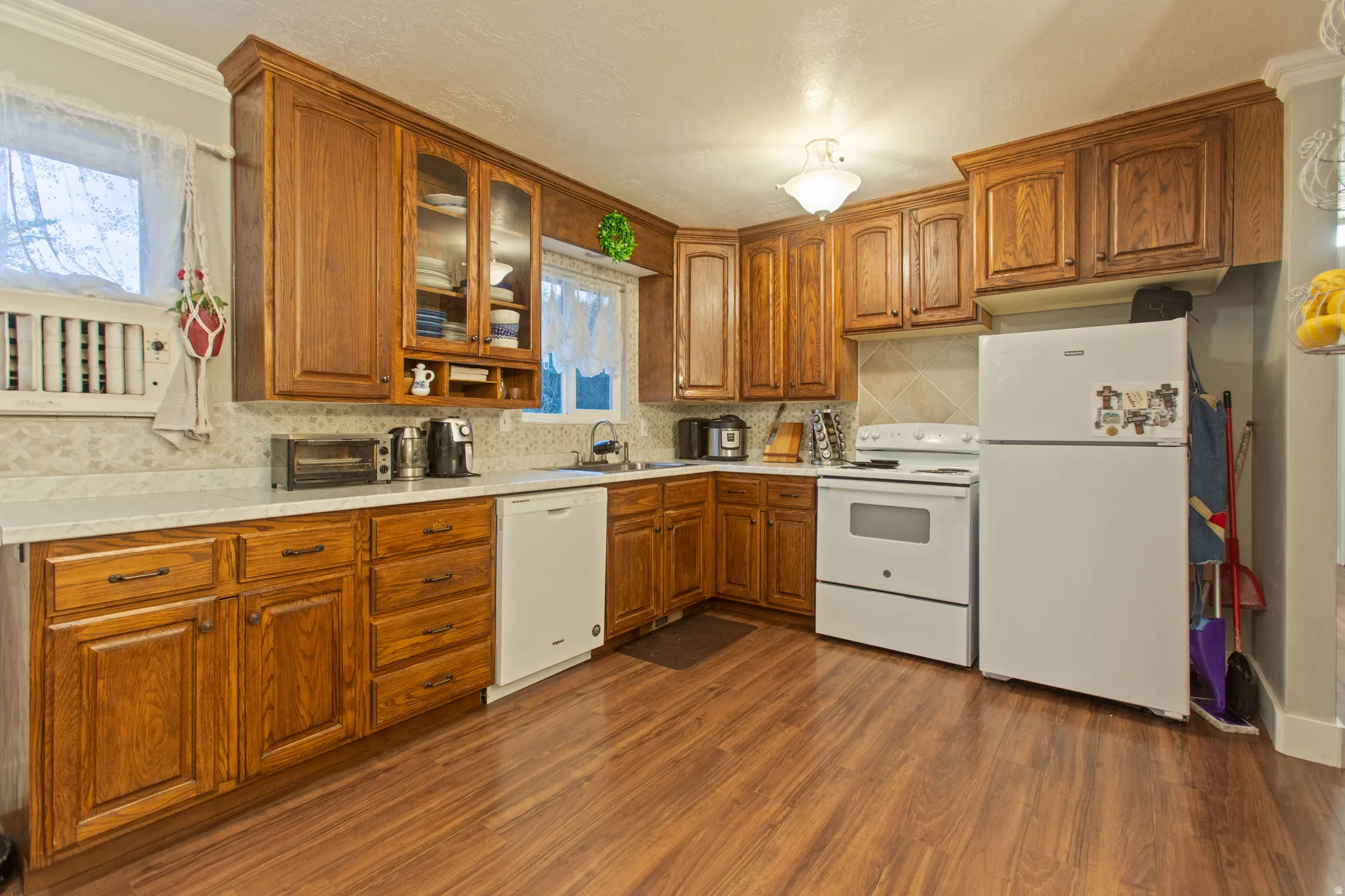 Wood-finish flooring, wood cabinetry with glass-front accents, white countertops, tile backsplash, and an integrated white dishwasher