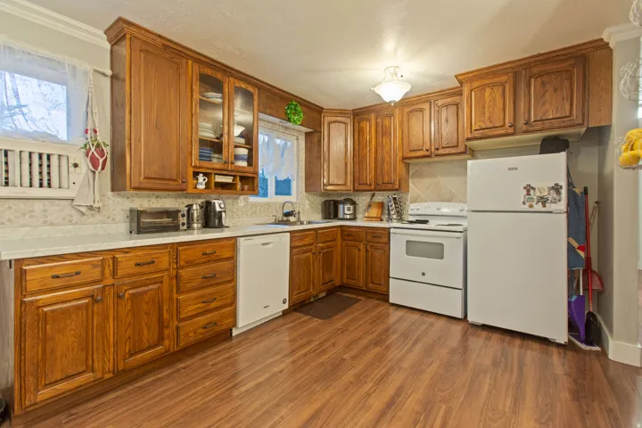 Wood-finish flooring, wood cabinetry with glass-front accents, white countertops, tile backsplash, and an integrated white dishwasher